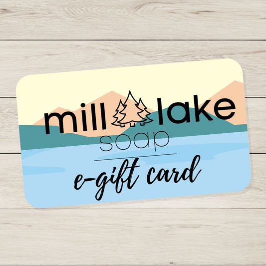 A digital gift card for Mill Lake Soap sits on a wooden table. The gift card has the Mill Lake Soap logo and black text that says "Mill Lake Soap e-gift card".